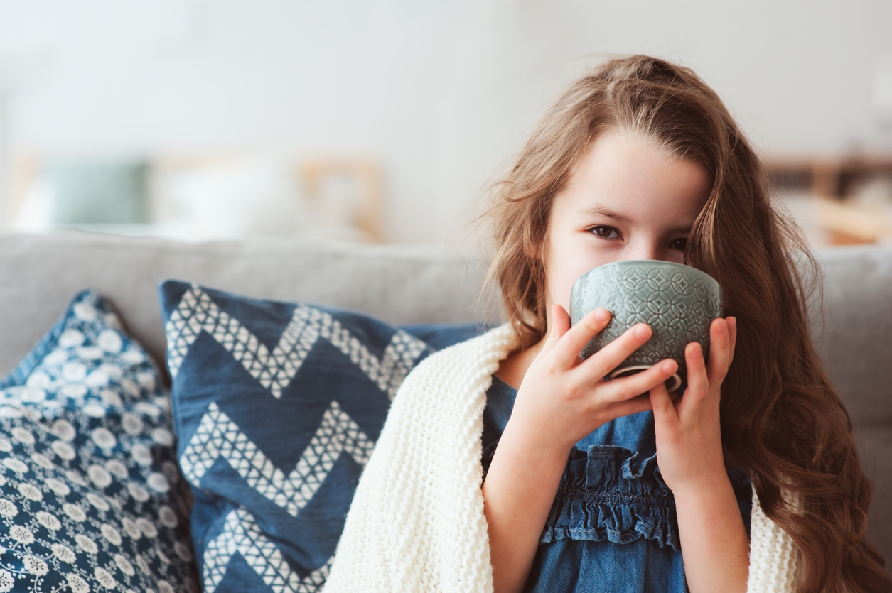 A little girl wrapped in a blanket drinking a warm drink because of a sore throat